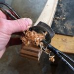 A spokeshave is pulled along the edge of a bent-laminate rocker, resulting in a handful of fine wood shavings.